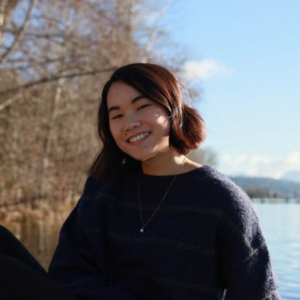 A person smiles outdoors near a body of water, with bare trees and blue sky in the background, suggesting a serene, natural setting.