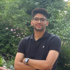 A person with glasses stands outdoors, wearing a black shirt and watch, amidst lush greenery and blooming flowers, looking at the camera.
