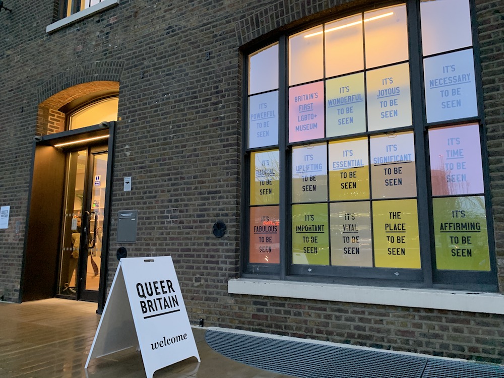 Brick building exterior with a sign reading "Queer Britain," showcasing Britain's first LGBTQ+ museum. Colorful window messages emphasize visibility and affirmation.