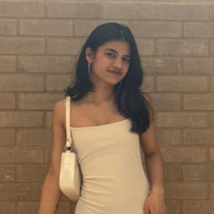 A person in a white dress stands against a brick wall, holding a white purse and smiling slightly, with long dark hair.