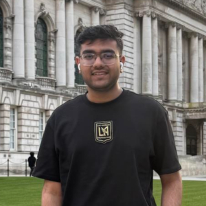 A person wearing glasses stands in front of a historical building with ornate columns, possibly Belfast City Hall, set against a grassy area.