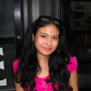 A person with long hair, wearing a pink top and sunglasses on head, smiles in front of a dark background indoors.