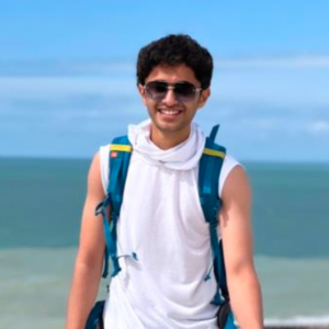 A person wearing sunglasses and a backpack stands smiling by a seaside, with a bright blue sky and ocean in the background.