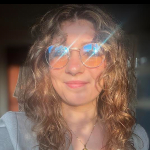 A person with curly hair smiles while wearing reflective glasses, catching sunlight. Warm lighting highlights the relaxed, casual atmosphere. No landmarks visible.