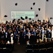 A jubilant group of graduates celebrate by tossing hats in the air at Richmond American University London.