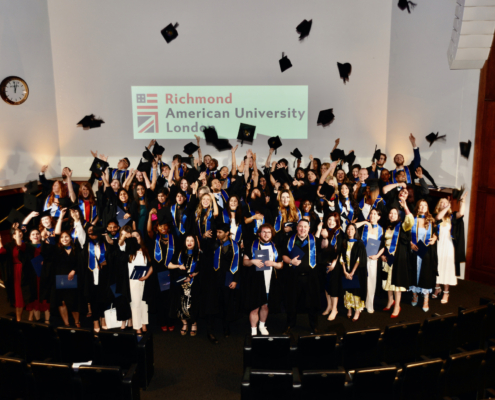 A jubilant group of graduates celebrate by tossing hats in the air at Richmond American University London.