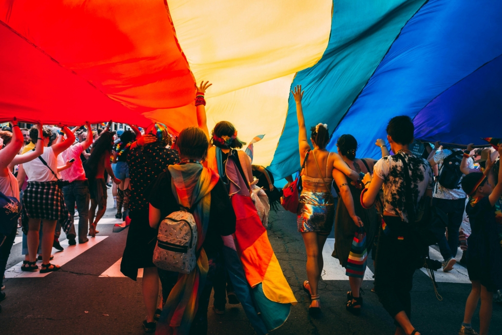People participate in a vibrant parade, holding a large rainbow flag overhead. The lively scene showcases diversity, joy, and community spirit.
