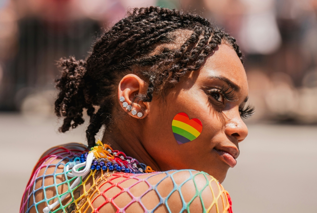 A person wears a rainbow heart sticker and colorful net top, showcasing pride, with a blurred background on a sunny day.
