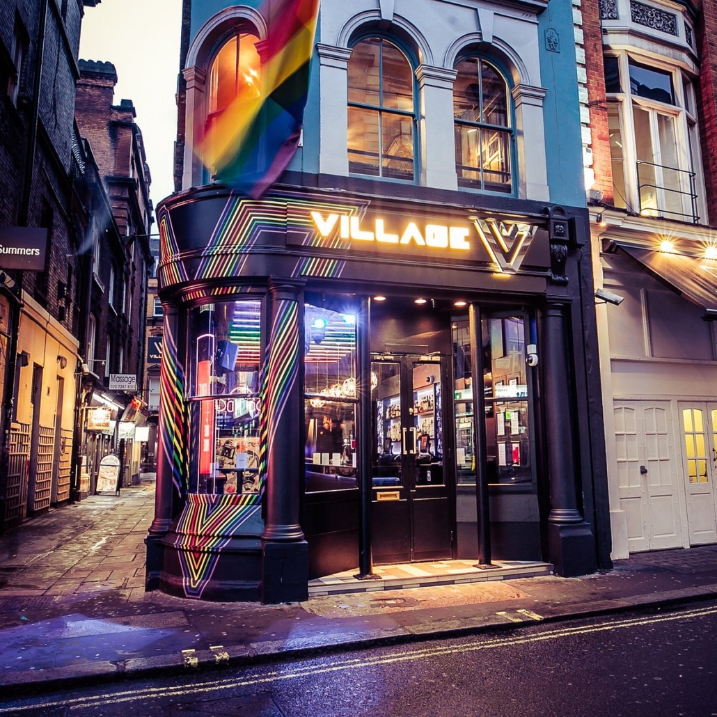 A vibrant bar with a rainbow flag in a narrow London street at dusk, featuring colorful neon lights and an inviting atmosphere.