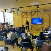 A person presents on "Businesses and Biodiversity" to a seated group in a modern, yellow-themed room with a television screen.