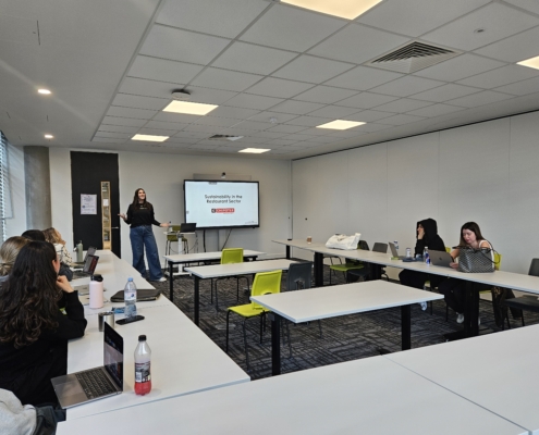 A person presents on sustainability in the restaurant sector to a small audience in a modern classroom with laptops and a projector.