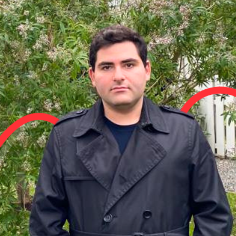 A person in a black coat stands outdoors against a background of leafy greenery and a white fence.
