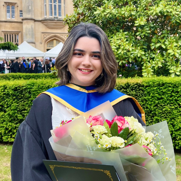 A person in graduation attire holds flowers, standing in front of historical building. Greenery and people in caps and gowns surround them.