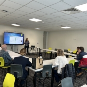 A person presents a restaurant value chain on a screen to seated individuals in a modern conference room with bright yellow chairs.