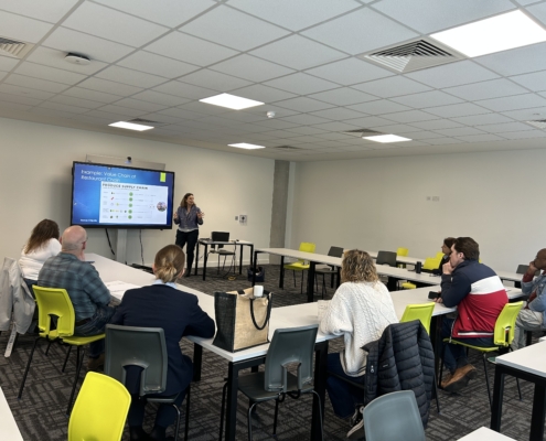 A person presents a restaurant value chain on a screen to seated individuals in a modern conference room with bright yellow chairs.
