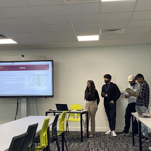 Four people present in a classroom, standing near a screen displaying a presentation. Tables, chairs, and laptops are arranged around them.