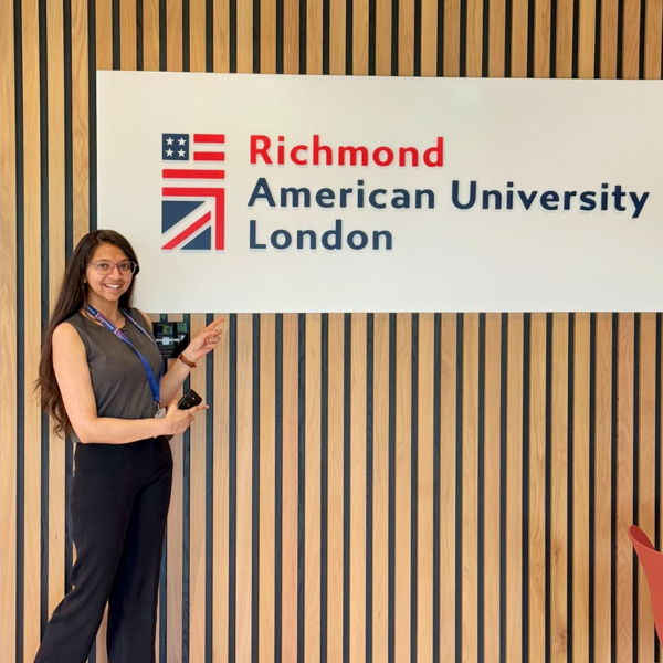 A person stands smiling, pointing at a Richmond American University London sign on a striped wooden wall.