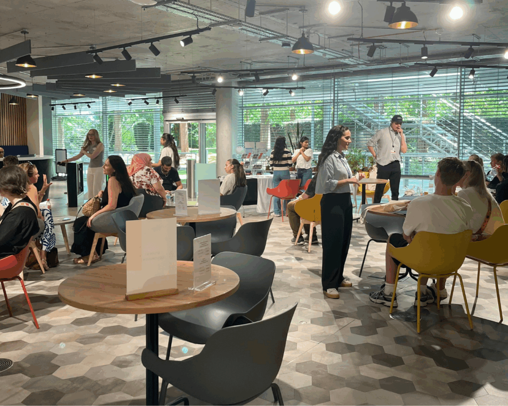 A modern cafe with several people socializing at tables. Natural light filters through large windows, creating a lively and inviting atmosphere.