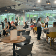 A modern cafe with several people socializing at tables. Natural light filters through large windows, creating a lively and inviting atmosphere.