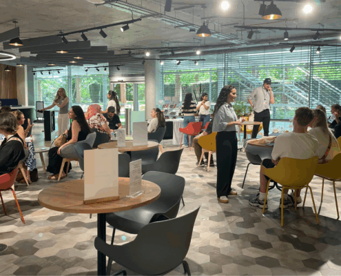 A modern cafe with several people socializing at tables. Natural light filters through large windows, creating a lively and inviting atmosphere.