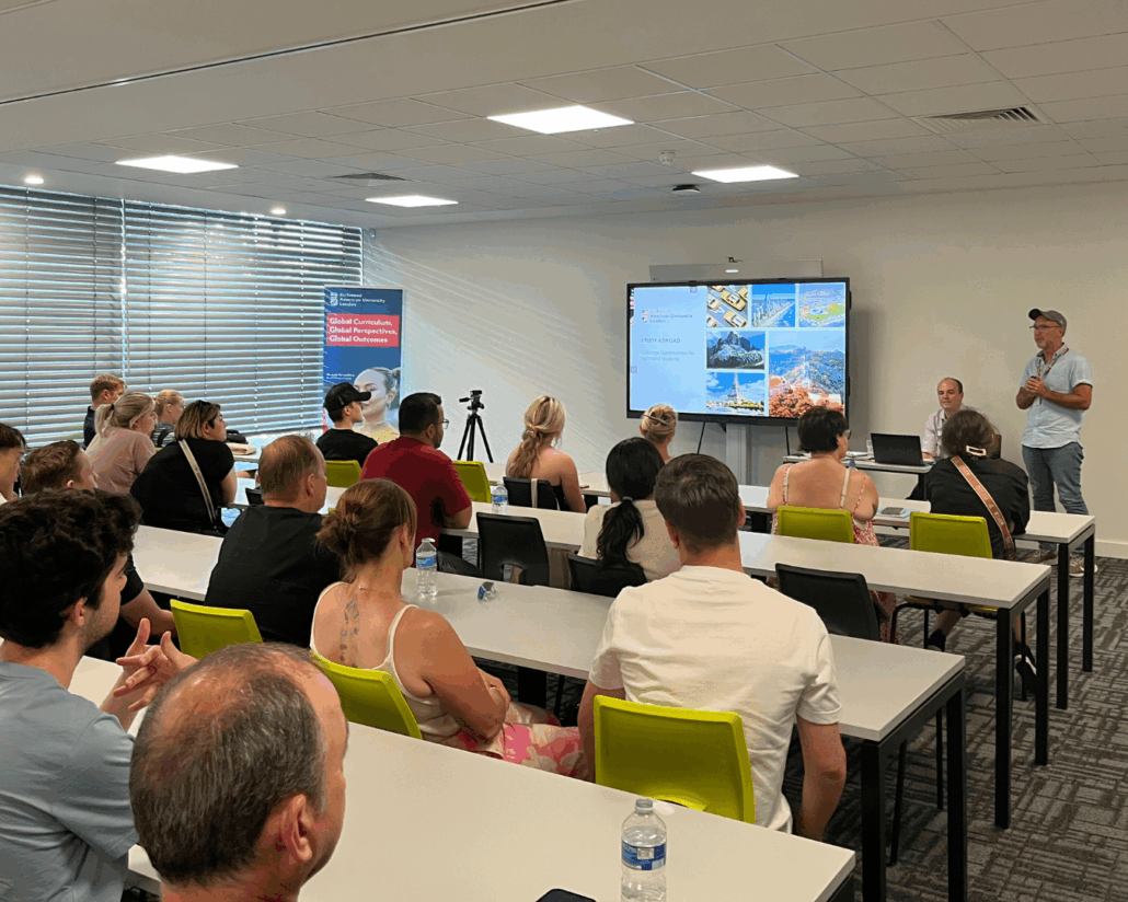 A classroom presentation with seated audience, a speaker, and slides displaying study abroad opportunities. Background includes informational posters and a camera.