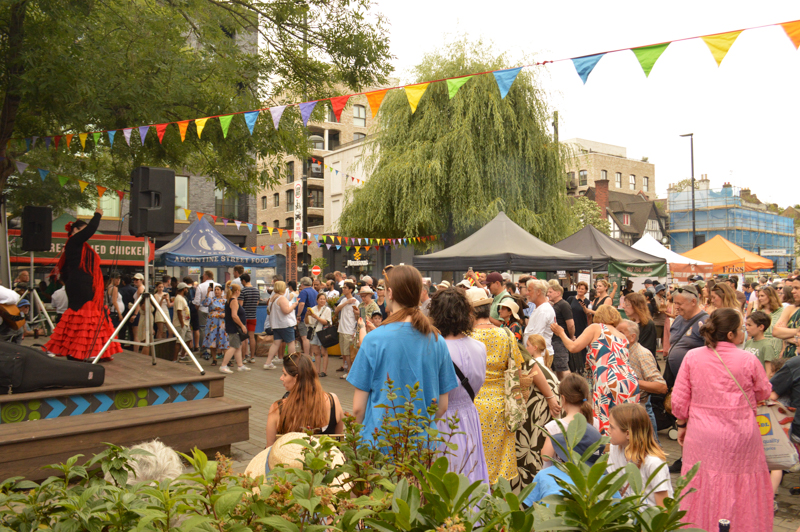 A vibrant street festival with bunting, diverse crowd, and a performer on stage. Food stalls and greenery enhance the lively atmosphere.