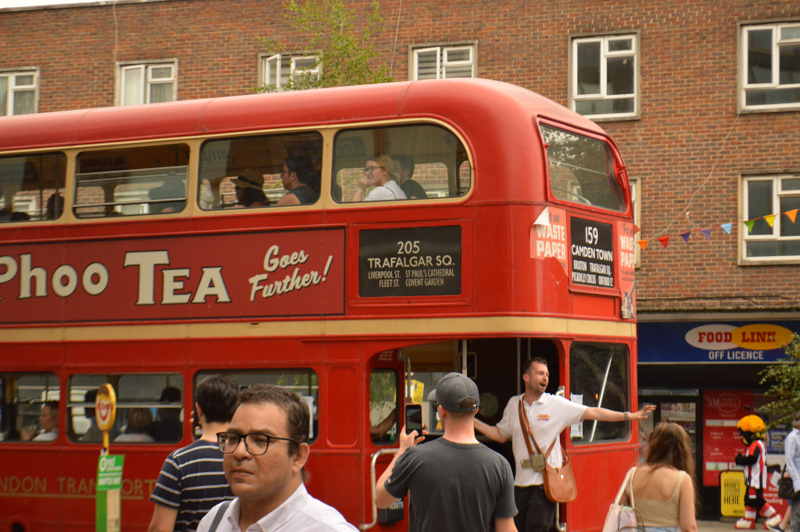 A red double-decker bus passes by people on a busy street with a brick building in the background; location suggests Trafalgar Square area.