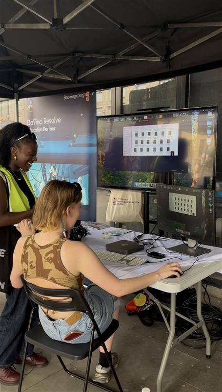 Two people at an outdoor booth, engaging with DaVinci Resolve software on screens. The setting includes promotional banners and computer equipment.
