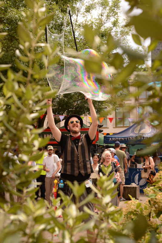 A person creates a giant bubble at a lively outdoor festival, surrounded by greenery and people enjoying the festive atmosphere.