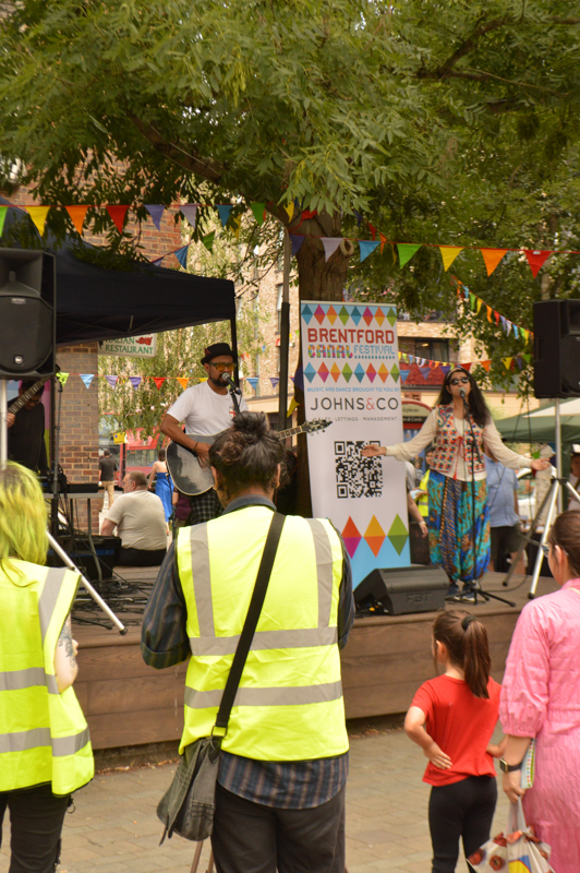 A lively outdoor performance at Brentford Canal Festival attended by people in yellow vests, with colorful bunting and a stage under trees.