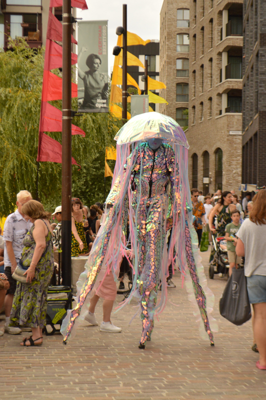 A crowd observes a person in a colorful costume resembling a jellyfish. Banners and brick buildings are visible in the background.