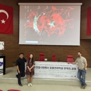 Three people stand in a conference room with Turkish flags, holding a banner. A large screen displays graphics above them.