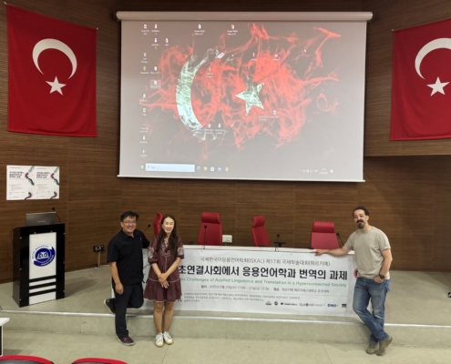 Three people stand in a conference room with Turkish flags, holding a banner. A large screen displays graphics above them.