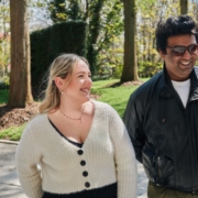 Two people smiling and walking in a park surrounded by trees. The scene is illuminated by bright daylight.