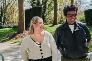Two people smiling and walking in a park surrounded by trees. The scene is illuminated by bright daylight.