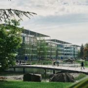 A scenic urban park features modern buildings, a pond, and a curved footbridge. People stroll and sit, enjoying the lush greenery.