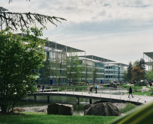 A scenic urban park features modern buildings, a pond, and a curved footbridge. People stroll and sit, enjoying the lush greenery.