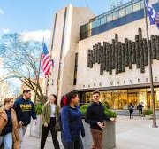 People walking in front of Pace University building, American flag visible. Sunny day, trees lining the street, university setting depicted.