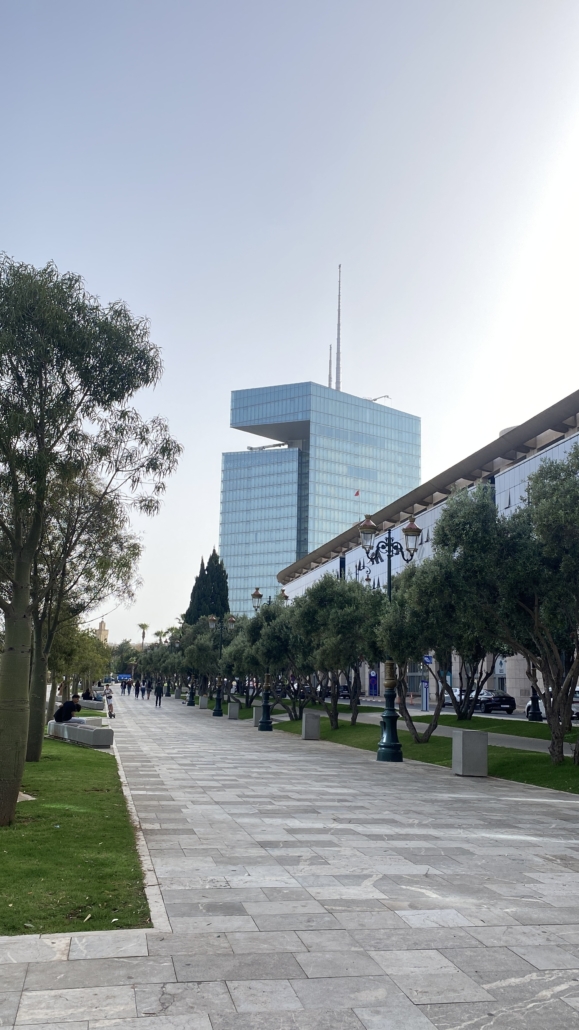 A modern glass building towers over a tree-lined walkway, where several people stroll and relax on benches on a bright day.