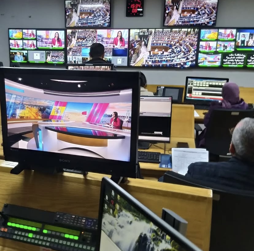 Control room with multiple screens displaying news broadcasts, people working at desks, and various live video feeds, including a news studio.
