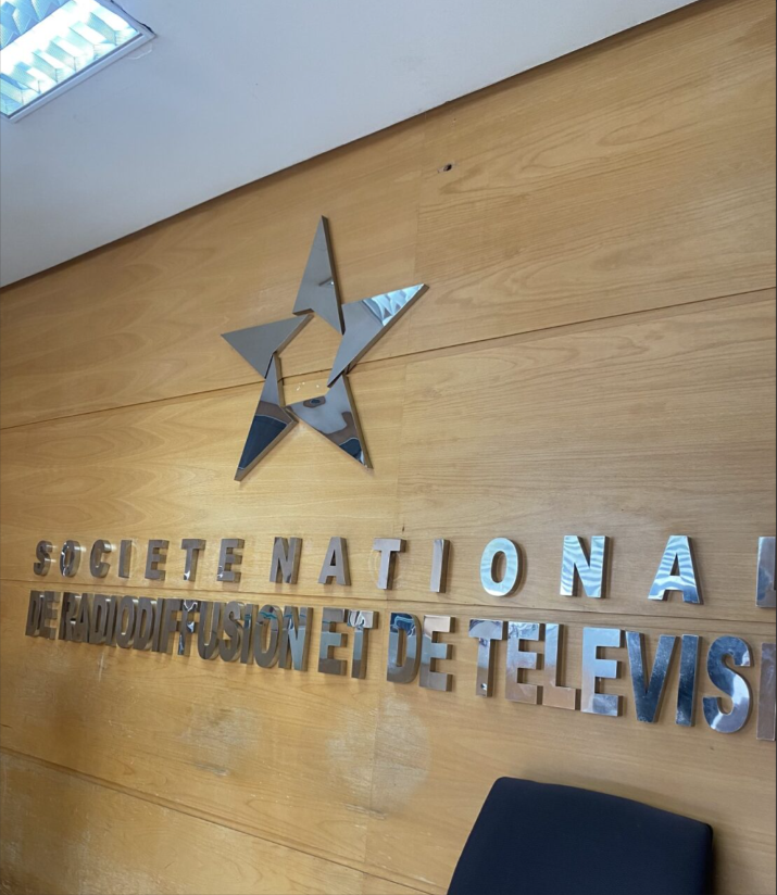 Wood-paneled wall with metallic star and text: "SOCIETE NATIONALE DE RADIODIFFUSION ET DE TELEVISION," modern design, interior setting, blue chair visible.