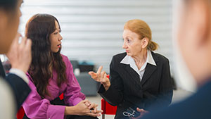 Two people engaged in conversation in a group setting, one gesturing expressively. Indoor environment with blurred individuals in the foreground.