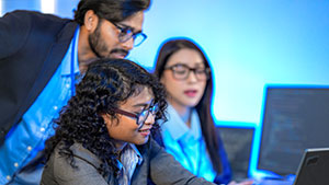 Three people in a modern office collaborate at computers, focusing intently. The room is illuminated by a blue hue, indicating a tech environment.