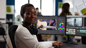 A person wearing headphones works at a computer with dual monitors, editing video. Blurred background shows another person in an office setting.
