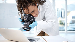 A person, wearing a lab coat and gloves, focuses a camera on a laptop in a bright, modern indoor setting.