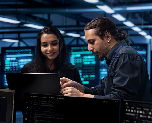 Two people are in a modern office, collaborating on a project with computers and digital screens displaying complex data.