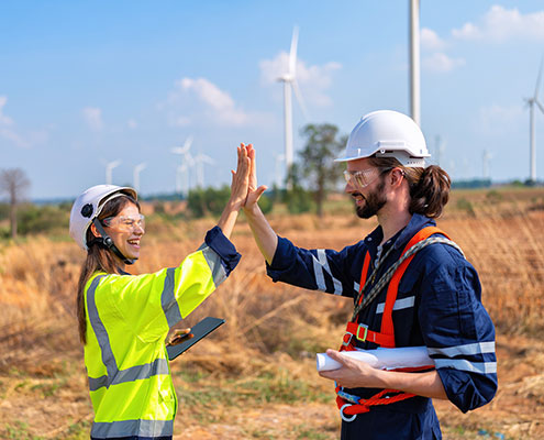 Two people in safety gear give a high-five near wind turbines in a grassy field, indicating teamwork and renewable energy efforts.