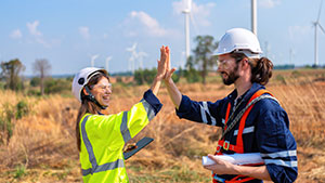 Two people in safety gear high-five in a field with wind turbines in the background, symbolizing teamwork and renewable energy development.