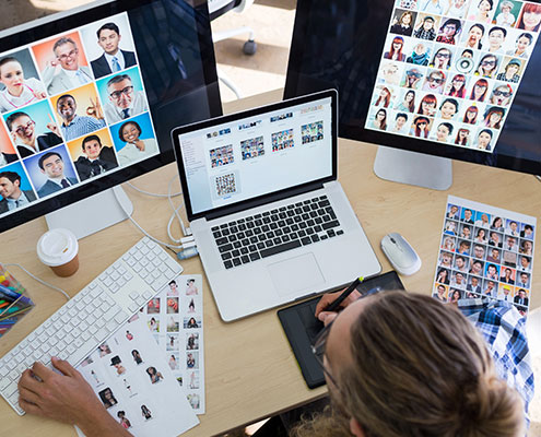 A person reviews multiple photos on computer monitors and printed sheets at a desk, likely organizing or selecting images for a project.