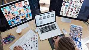 A person works at a desk with three monitors displaying various people’s photos, surrounded by printed images, pencils, and a coffee cup.
